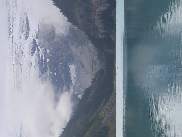 A turquoise glacial lake surrounded by steep forested slopes and mist-covered snowy peaks rising into the clouds.