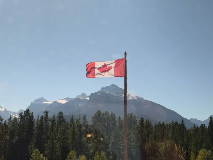 A Canadian flag waves on a tall pole against a backdrop of evergreen forest and snow-capped Rocky Mountain peaks under a clear blue sky.