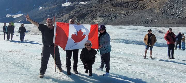 Travellers celebrate on a glacier holding a large Canadian flag while other visitors explore the icy terrain nearby.