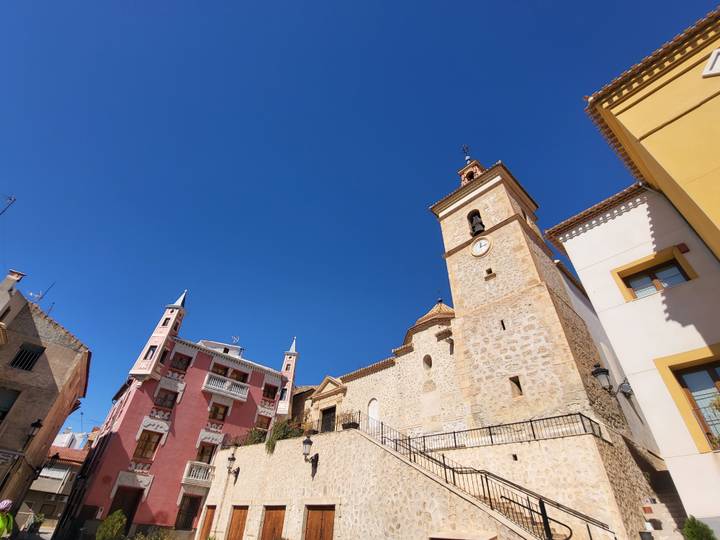 Upward view of a stone church tower with clock next to colorful historic buildings under a deep blue sky in Cieza.