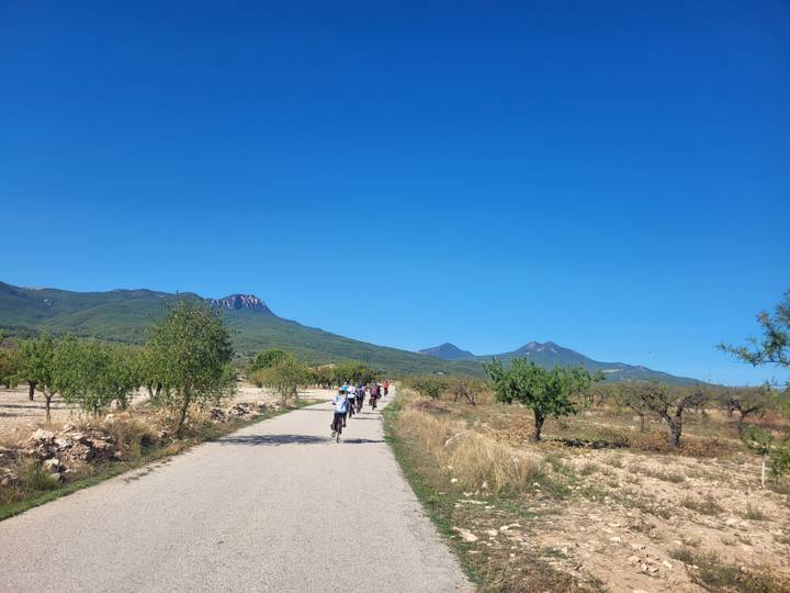 Cyclists ride along a rural path flanked by almond trees with mountain ranges under a clear blue sky.