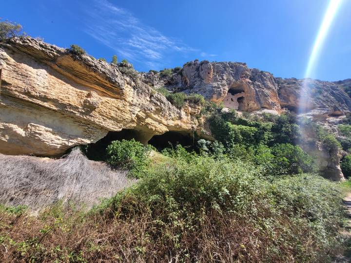 Rocky cliffside with natural caves and lush vegetation illuminated by a shaft of sunlight.