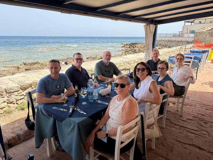 Travel party shares a meal by the sea on a sunny Sicilian afternoon with colorful chairs and stone promenade.