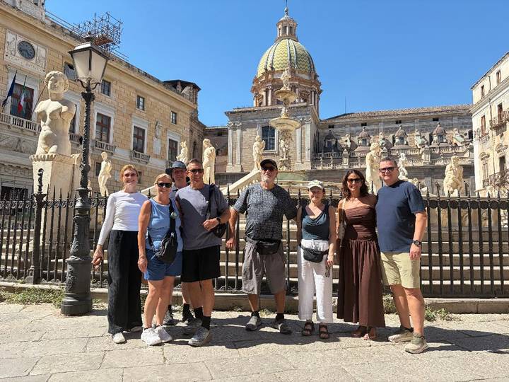 Group stands before the ornate Fontana Pretoria and domed church in historic Palermo on a bright day.