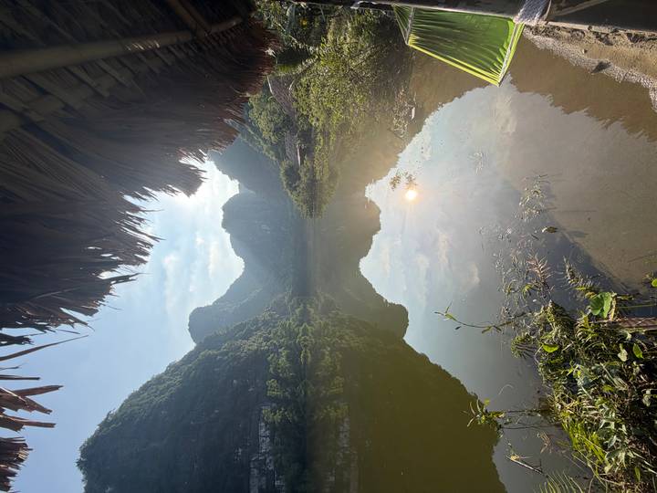 Serene lake mirroring karst mountains and morning sun seen from a thatched riverside hut.