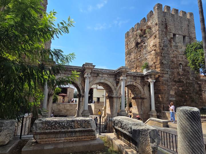 Roman-era stone arches and towers of Hadrian's Gate with tourists exploring the historic site.