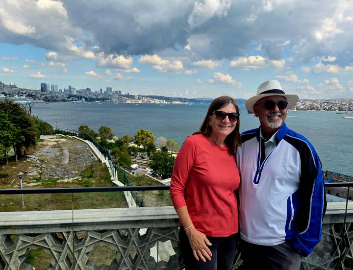 Couple stand by a railing overlooking the Bosphorus with Istanbul skyline in the distance under dramatic clouds.