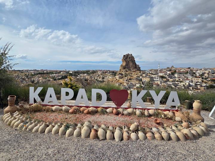 Large sign spelling KAPADOKYA with heart symbol in front of Uchisar Castle and Cappadocia townscape.