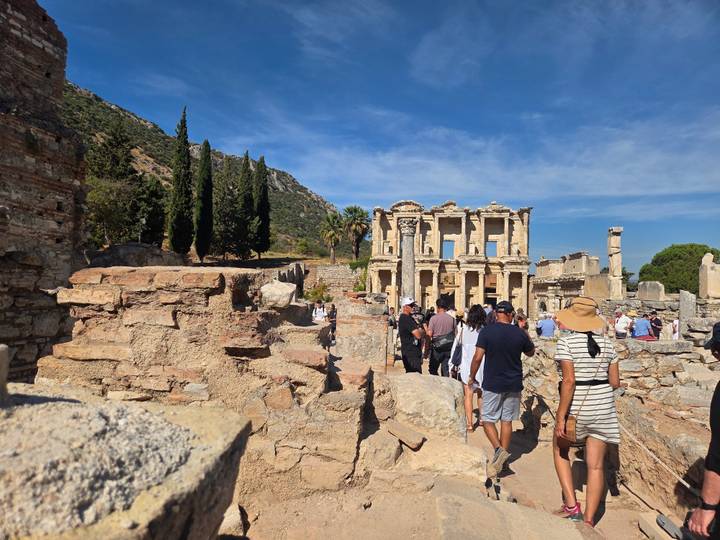 Tour group walks among the ruined walls and columns of ancient Ephesus under bright sky.