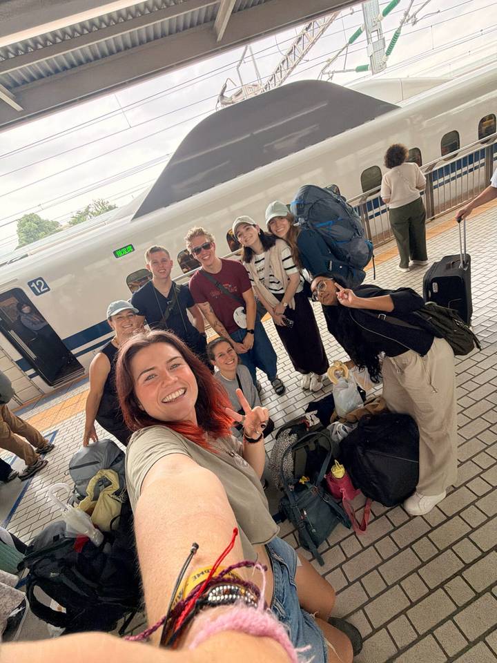 Smiling group selfie on a Shinkansen platform with backpacks and a high-speed train behind.