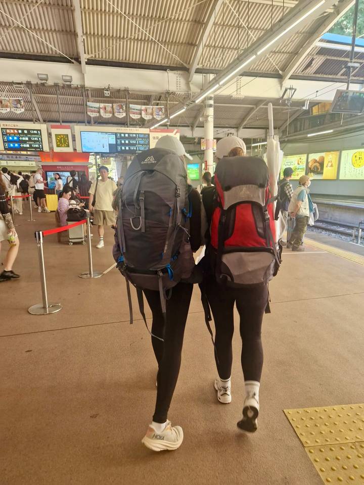Two backpackers standing side by side on a busy Japanese train platform.