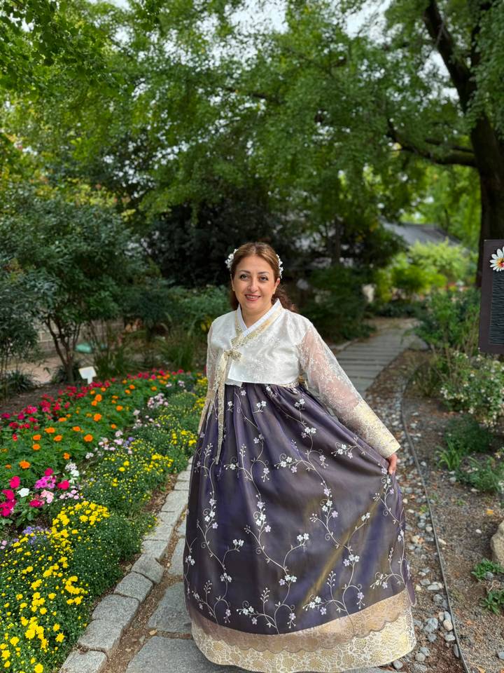 A woman in a purple floral hanbok twirls happily in a colourful garden lined with blooming flowers and shady trees.