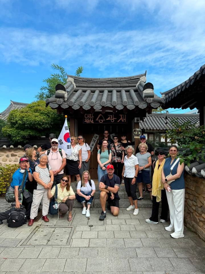 A large tour group poses outside a traditional Korean wooden gate, holding a South Korean flag beneath a clear blue sky.