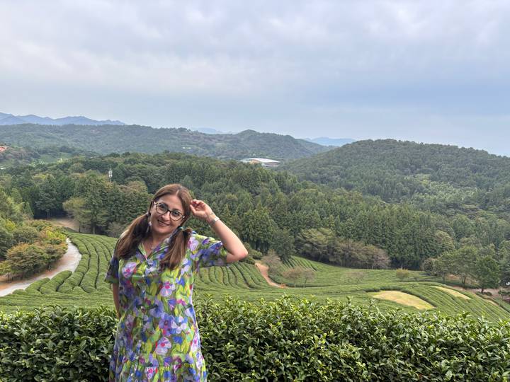 A traveller poses playfully before rolling green tea plantations stretching across misty hills under a soft blue sky.