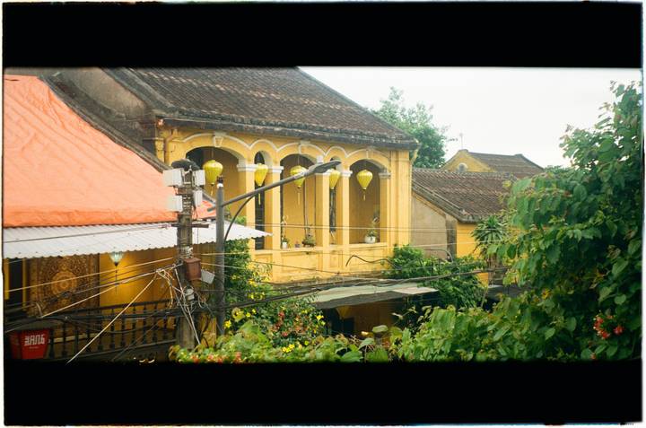 Yellow colonial-style house with arched balconies and tiled roofs framed by lush vegetation and overhead power lines.