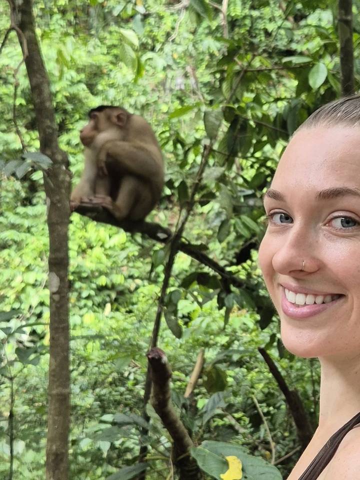 Close-up selfie of a smiling traveller with a monkey perched on a branch in the blurred jungle background.