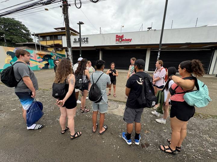 Tour group gathers around a guide on a graffiti-lined urban street with shuttered storefronts.