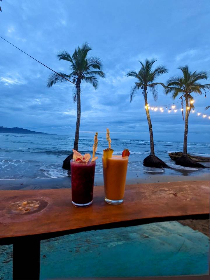 Two colorful cocktails rest on a seaside bar counter with palm trees, string lights, and a twilight ocean backdrop.