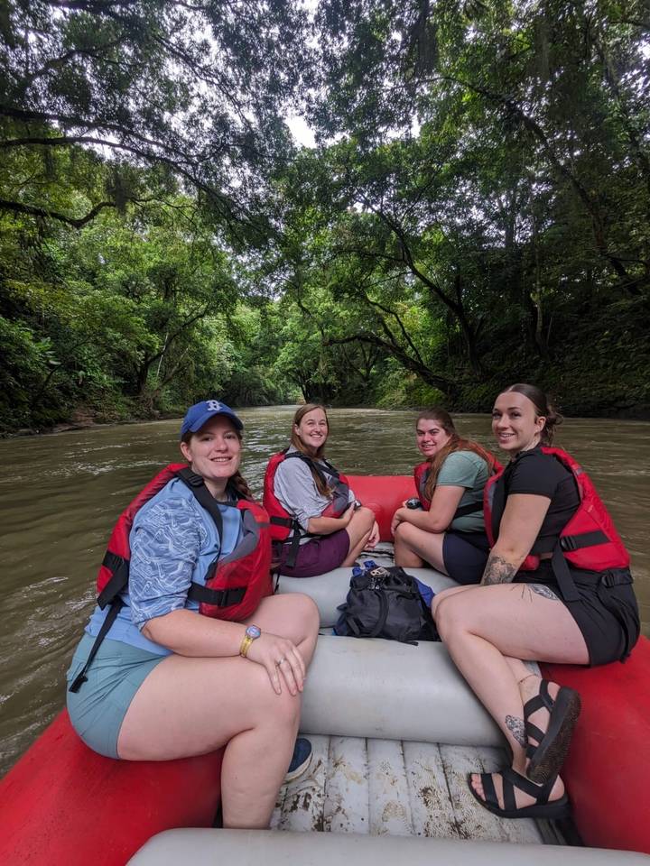 Group of friends wearing life jackets sit in an inflatable raft floating through a jungle river canyon.