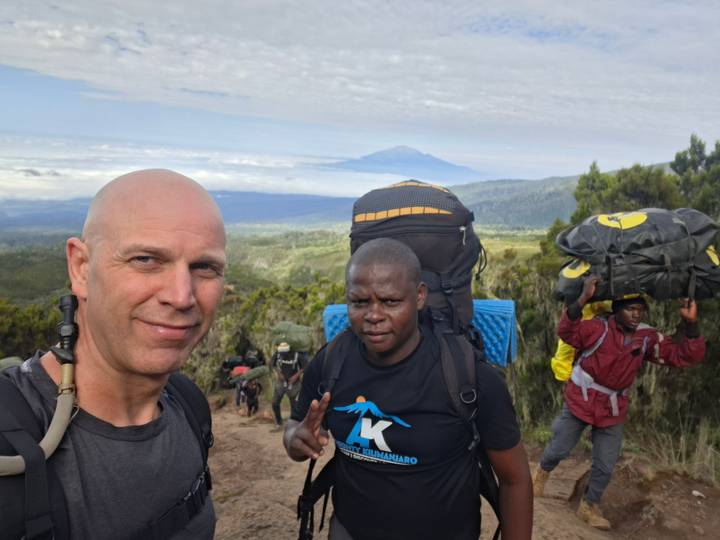 Hikers posing on a mountain trail with backpacks while porters carry gear and Mount Kilimanjaro rises in the distance.