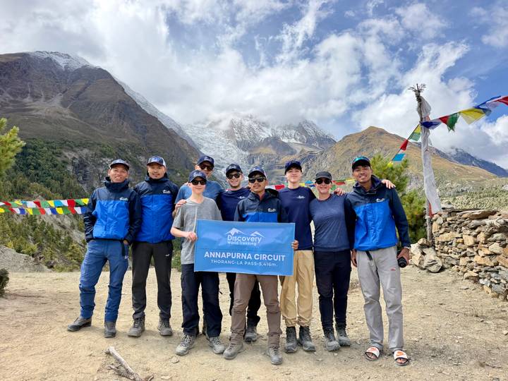 A trekking group celebrates at a high-altitude pass holding an Annapurna Circuit banner with rugged peaks behind.