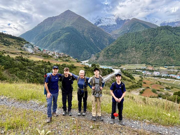 A family of trekkers smiles on a grassy ridge overlooking a broad Himalayan valley dotted with villages.