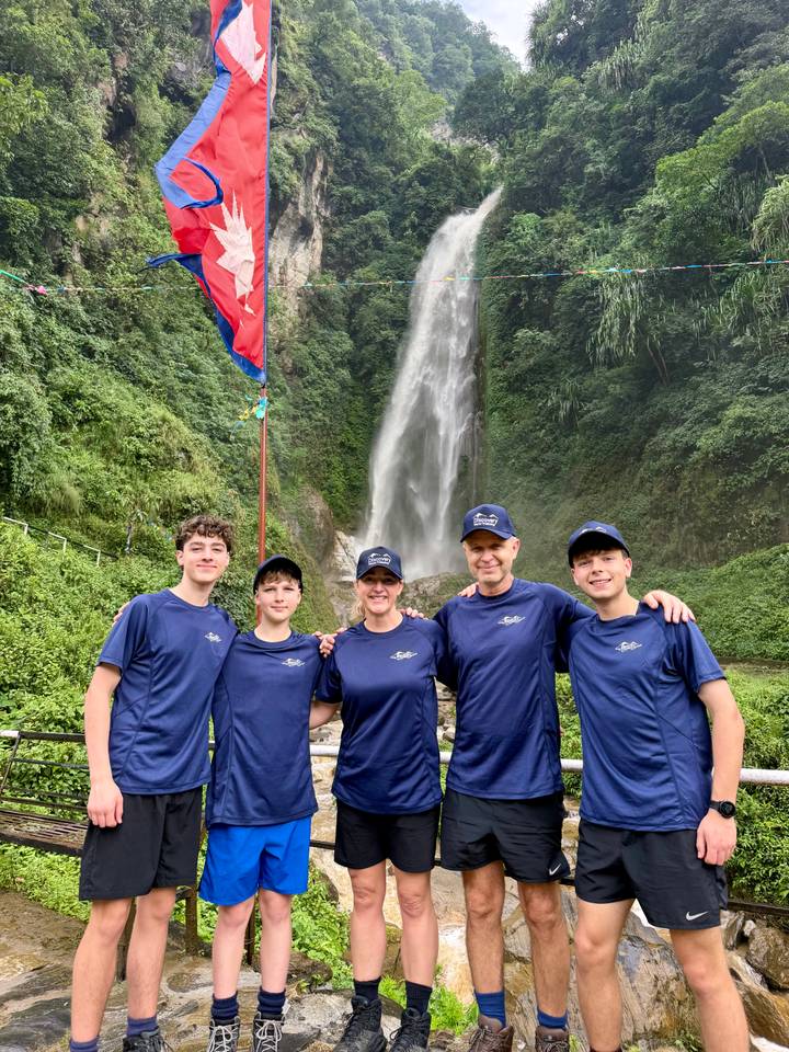 A smiling group stands arm-in-arm before a tall rainforest waterfall, Nepalese flag fluttering beside them.