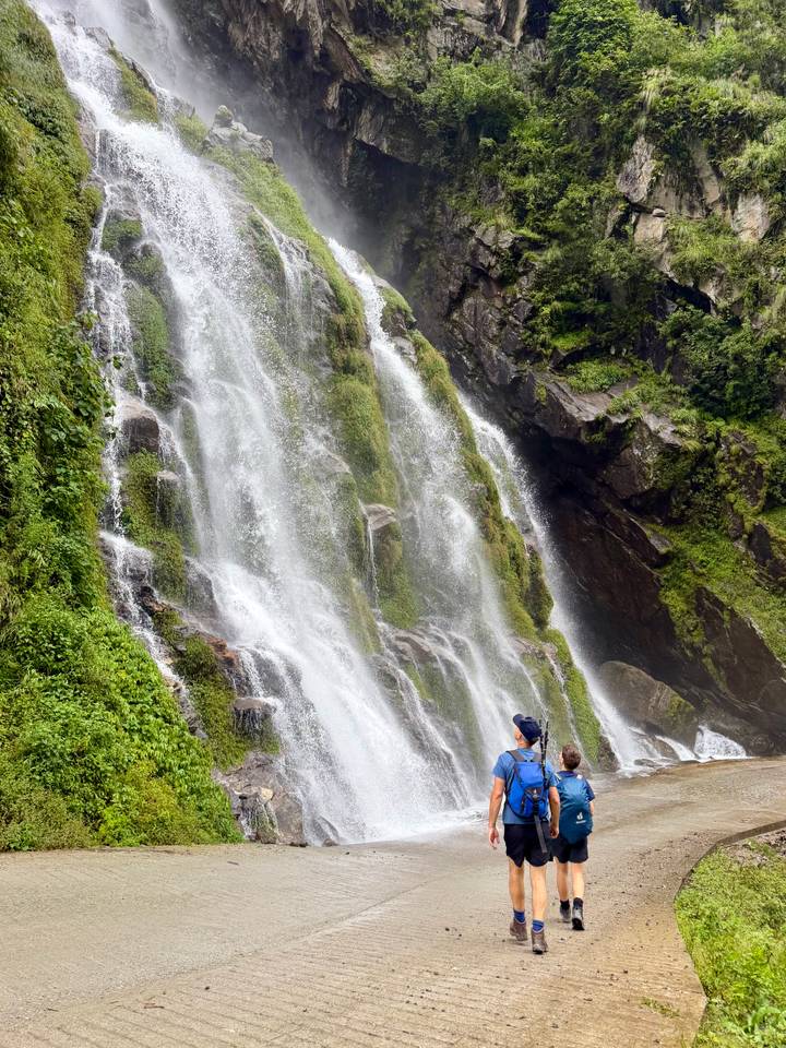 A tall, multi-tiered waterfall plunges down a mossy cliff while a lone hiker gazes up from the base.