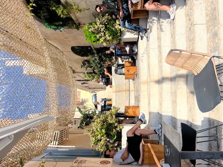 Sunny stone alley with steps lined by plants and café tables, mesh canopy casting patterns overhead