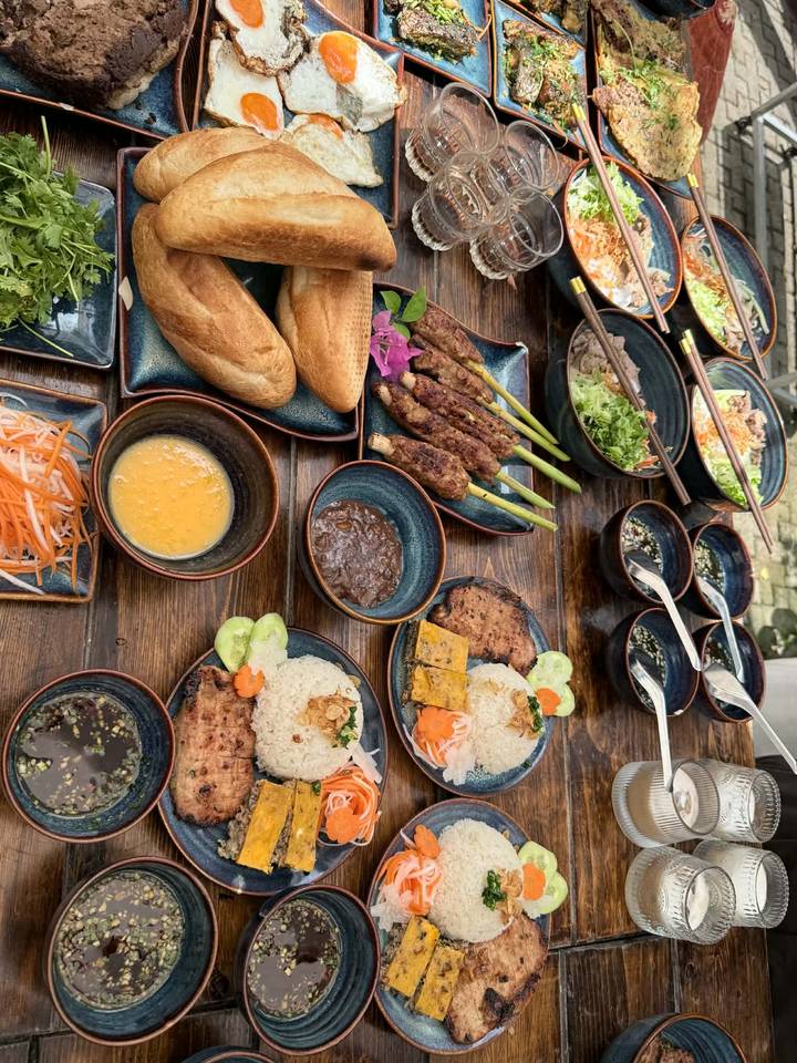 Overhead spread of Vietnamese dishes including grilled pork, baguettes, rice, pickles, and dipping sauces on a rustic table.