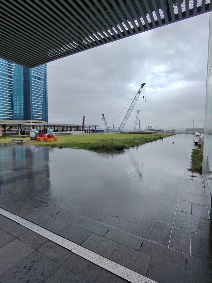 Rain-soaked plaza by a construction site with scattered sculptures under grey skies in Busan.
