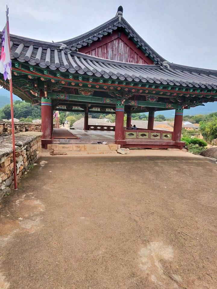 Traditional wooden pavilion in rural Korea with visitors resting on raised platform.