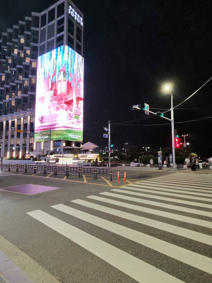Night intersection lit by giant LED adverts and crosswalk near Busan beachfront.