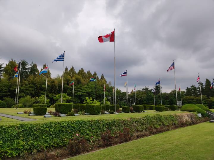 Line of international flags fluttering above manicured hedges under brooding clouds at memorial grounds.