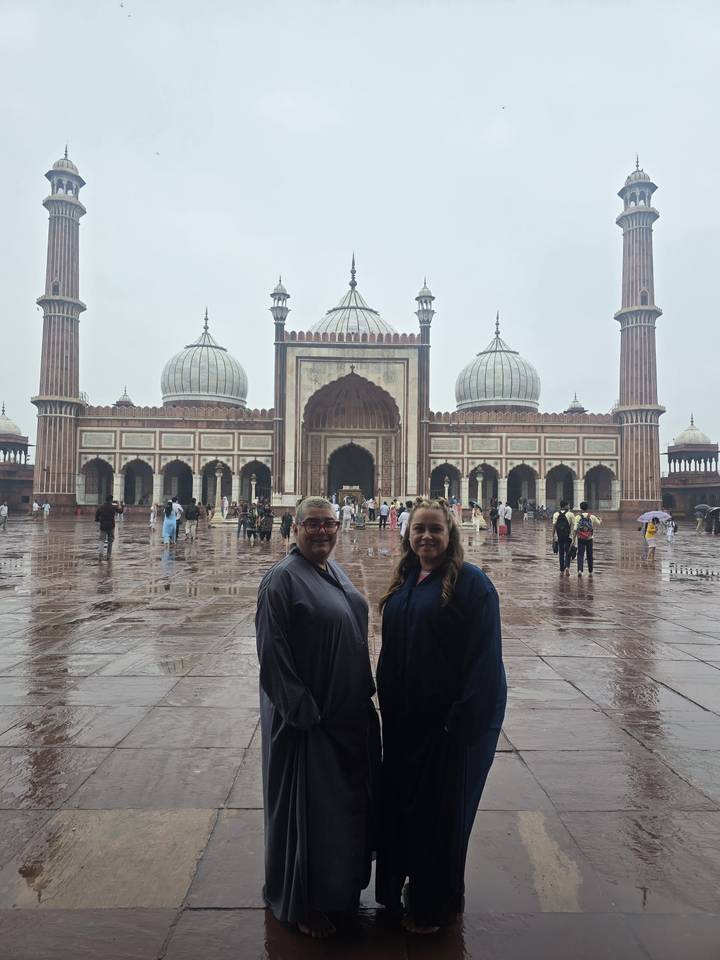 Two visitors in modest attire stand on the wet marble courtyard of Jama Masjid with its domes and minarets rising behind.