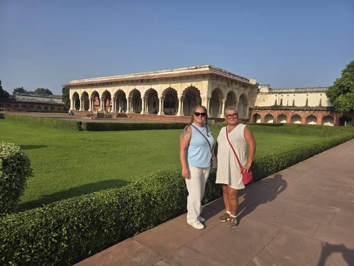 Visitors stand in a sunny green courtyard framed by ornate arches of an historic red-sandstone fort.