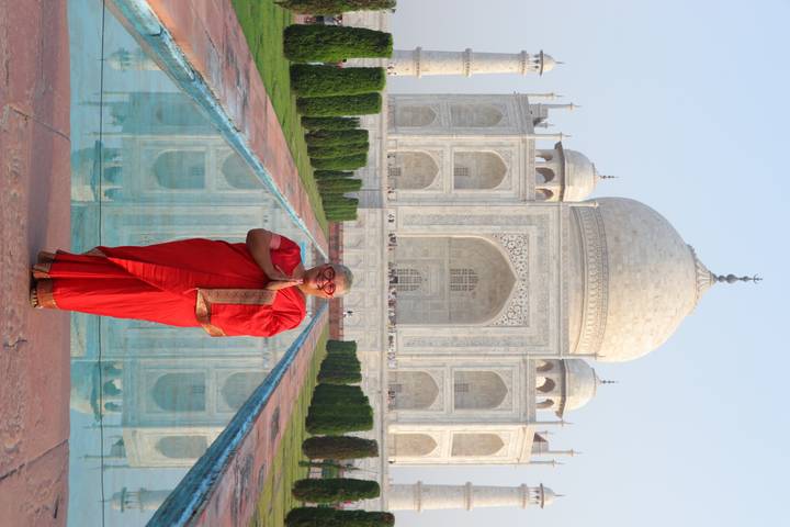 Woman in red sari stands with hands pressed in namaste before the reflecting pool and Taj Mahal.
