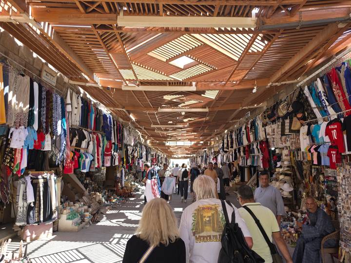 Covered souk lined with stalls selling colourful clothing and souvenirs, busy with shoppers.