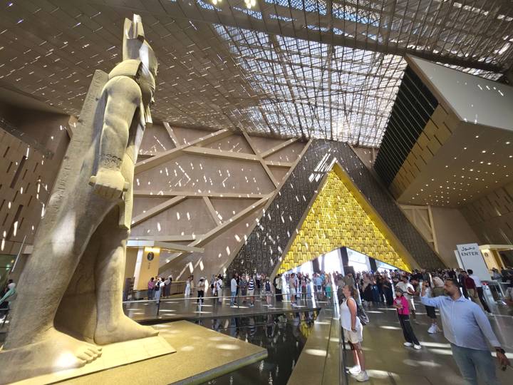 Grand Egyptian Museum atrium with modern geometric ceiling and colossal statue drawing crowds of visitors.