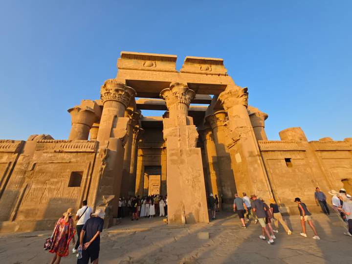 Golden-hour view of Kom Ombo Temple’s massive pillars and carved reliefs with tourists gathering at the entrance.
