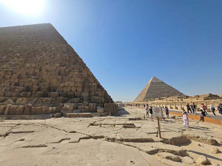 Large stone pyramids loom over visitors walking the Giza Plateau on a bright, clear day.