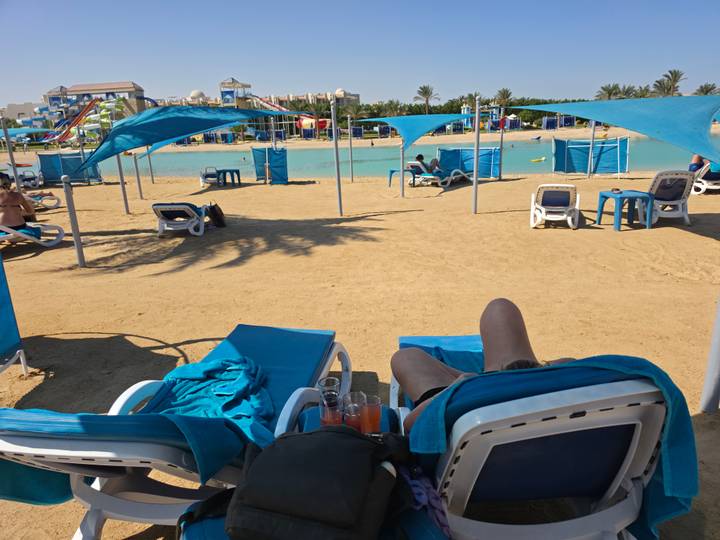 Relaxing beach club scene with sun loungers and shade sails on golden sand beside a turquoise lagoon.