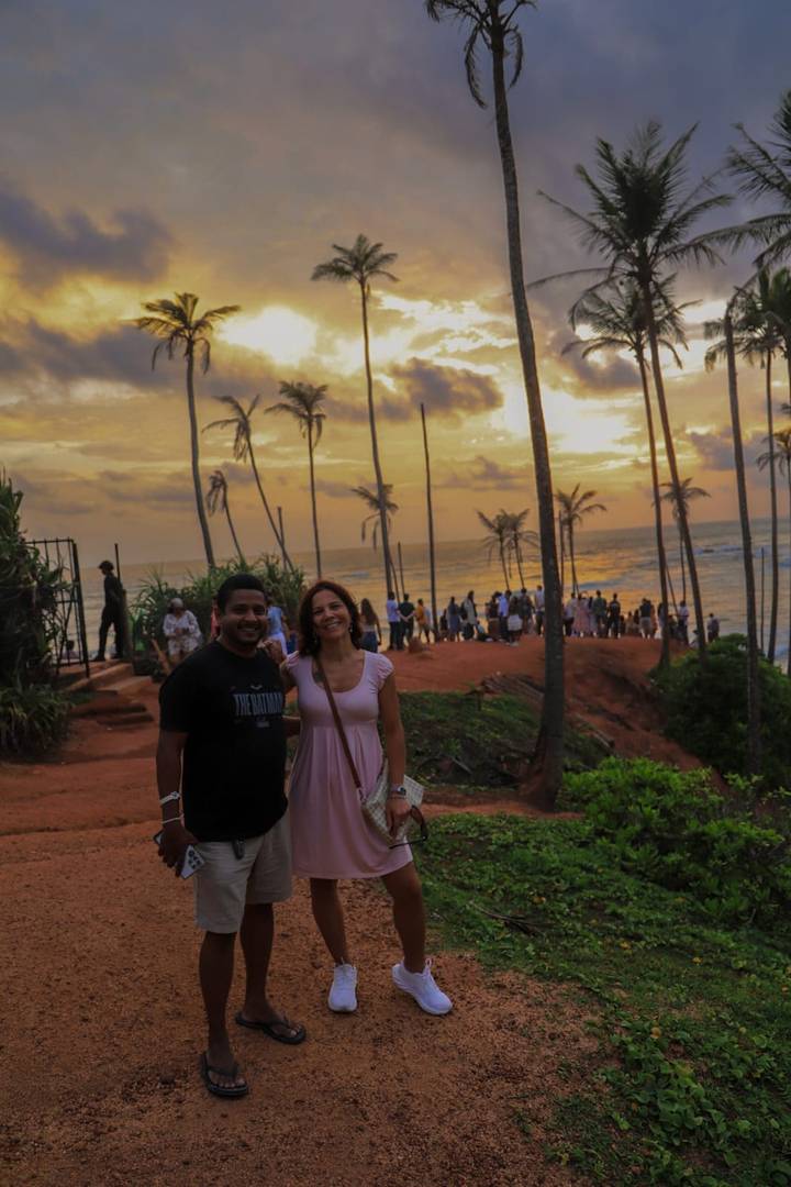 Couple smiles at Coconut Tree Hill with crowds gathering to watch the sunset over the ocean.