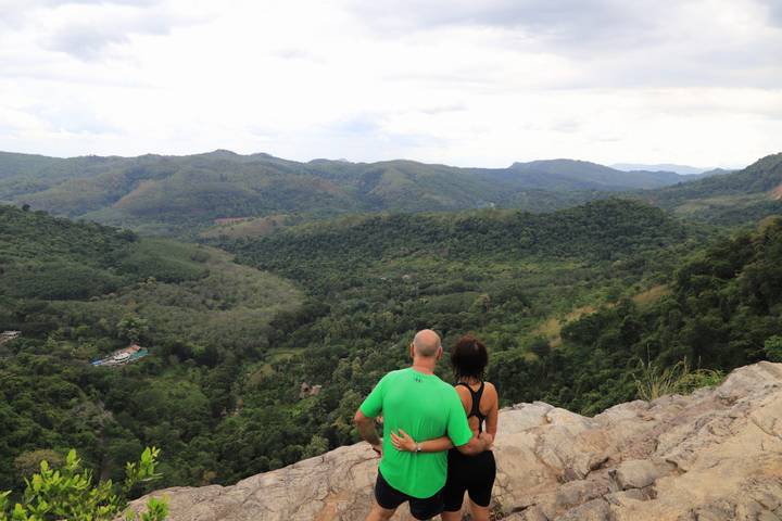 Couple sits on rocky outcrop admiring a vast green valley and distant mountains.