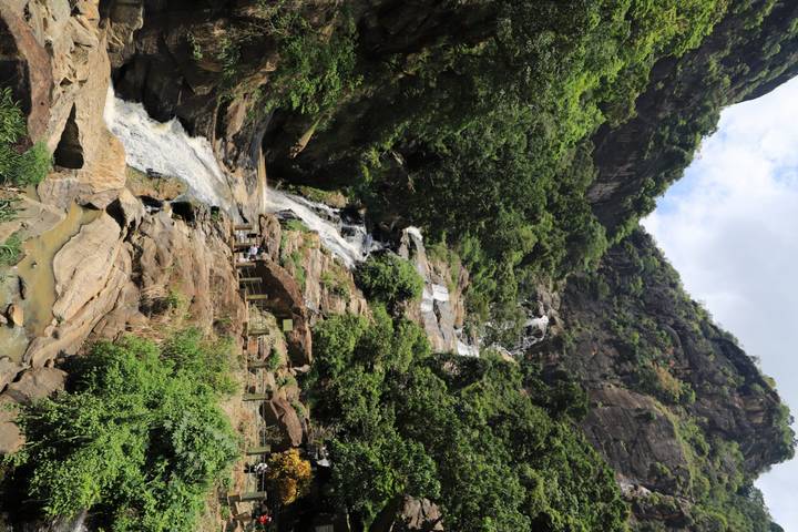Tall multi-tier waterfall cascades down a lush cliffside framed by tropical foliage.