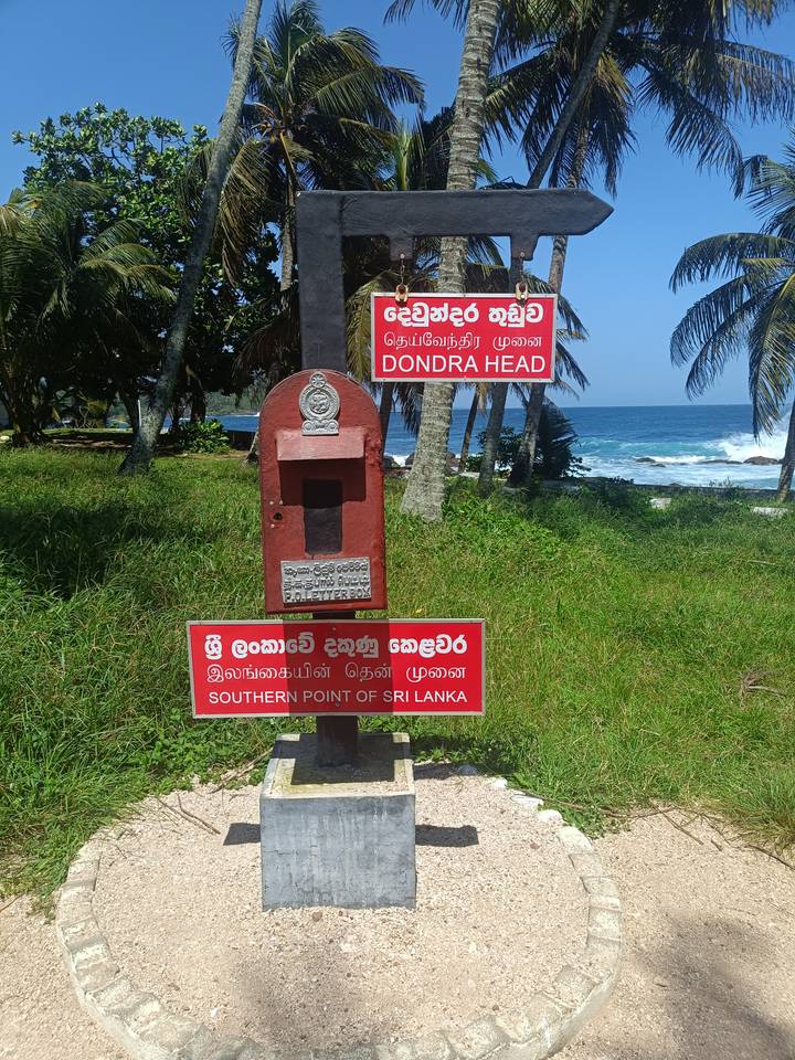 Red signposts mark Dondra Head, the southernmost point of Sri Lanka, with palm trees and turquoise sea beyond.