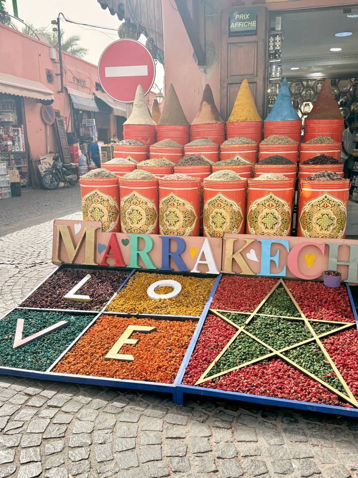 Colorful spice barrels arranged around a large “Marrakech” sign in a market.