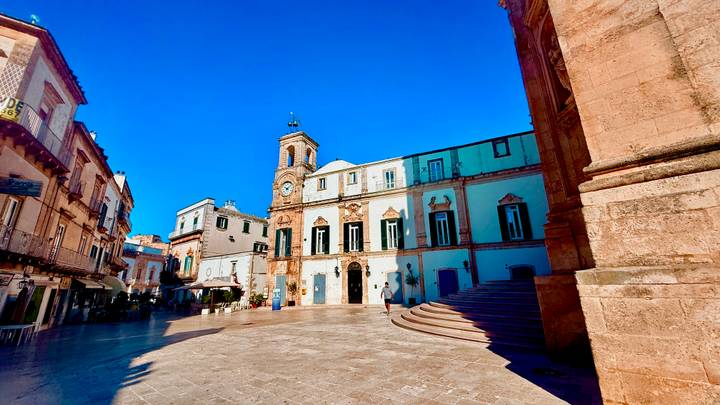 An elegant Italian piazza with clock tower, pastel buildings and wide stone pavement.