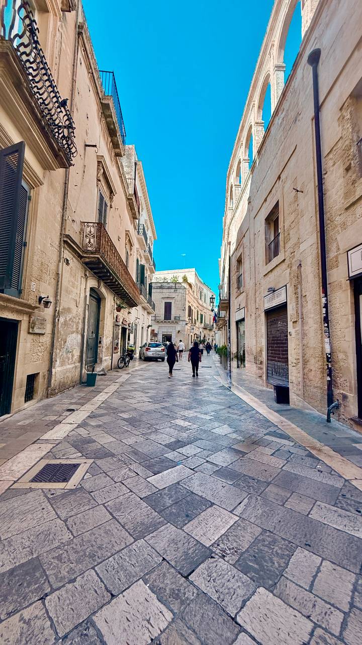 Pedestrians walk a narrow stone street lined with historic limestone buildings.