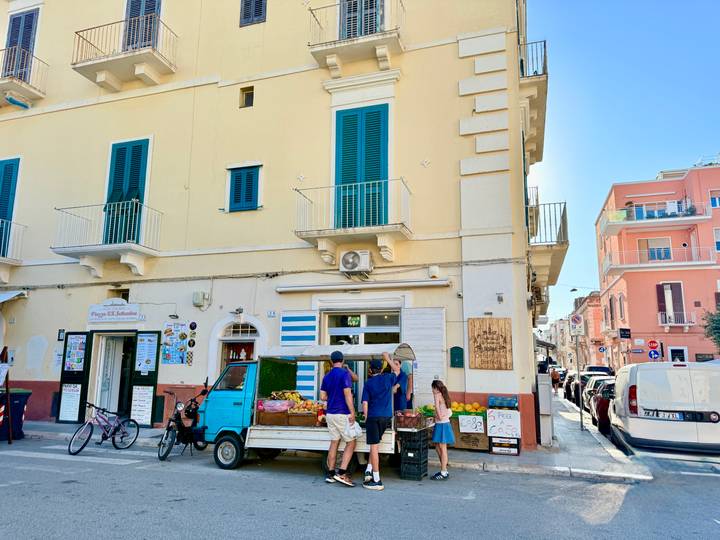 Locals buy produce from a small blue three-wheeled truck outside pastel buildings.